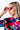 A person with blonde hair adjusts their BAT WING SUNNIES, which are pink and angular. They are wearing a vibrant shirt with a shiny texture, featuring an abstract rainbow-colored pattern. The background is plain white.