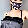 A woman with long dark hair wears a black dress with spaghetti straps and gold buttons. She has large hoop earrings and the BE BOLD FASHION FACE COVER, a black face mask featuring white, bold, graphic text. She stands against a plain, light-colored background, looking directly at the camera.