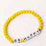 The BE HAPPY BRACELET features yellow beads and a section of white beads that spell out "BE HAPPY" in black letters. The design is accentuated with two gold beads placed on either side of the white beads, and it is showcased on a plain white surface.