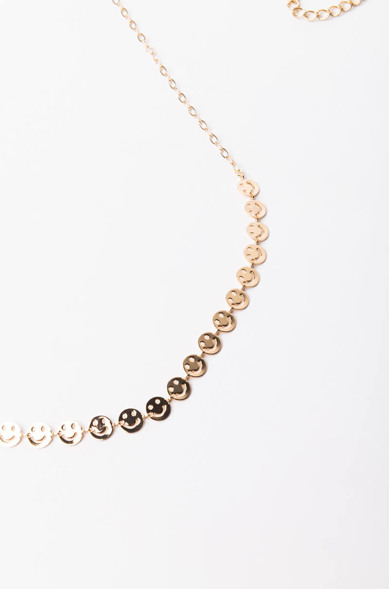 The BECAUSE I'M HAPPY BELLY CHAIN is a minimalist gold accessory adorned with a series of small, circular smiley face charms linked in sequence. The chain extends from both sides of the smiley face arrangement, creating a striking contrast against a plain white background.
