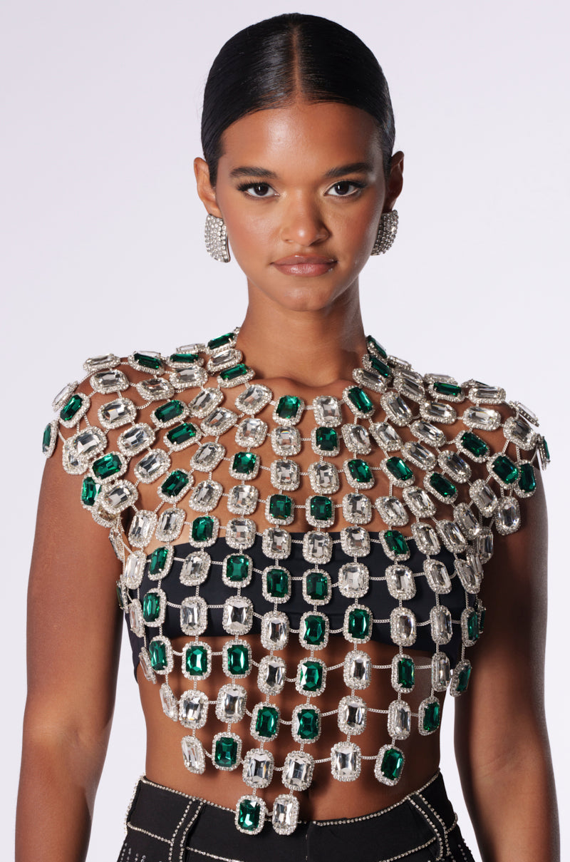 A woman with dark hair tied back is wearing a BEJEWLED EMERALD SLEEVELESS TOP, embellished with large, rectangular green and clear gemstones. The top showcases intricate patterns on a black base. She complements the outfit with matching earrings against a plain white background.