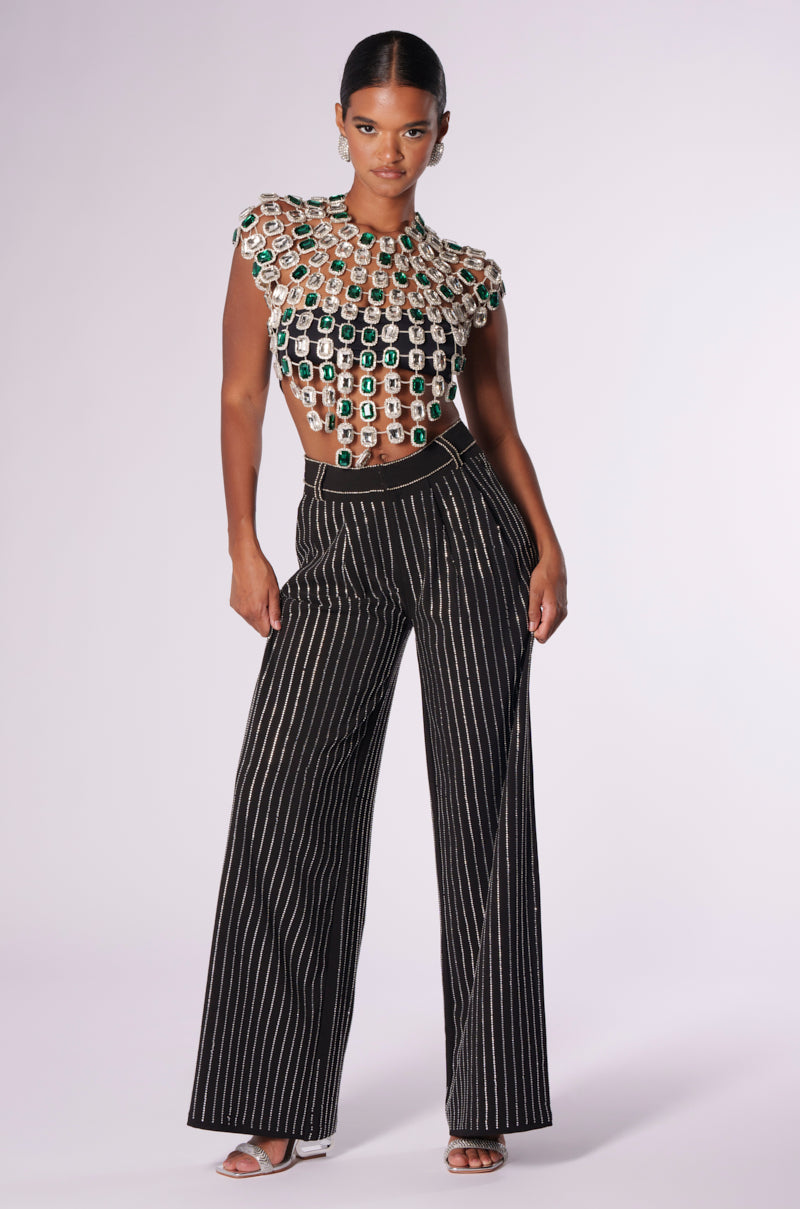 A model poses in a stylish outfit, featuring the BEJEWELED EMERALD SLEEVELESS TOP adorned with intricate gemstone embellishments, paired with high-waisted, wide-leg striped pants. She stands against a plain background, exuding confidence.