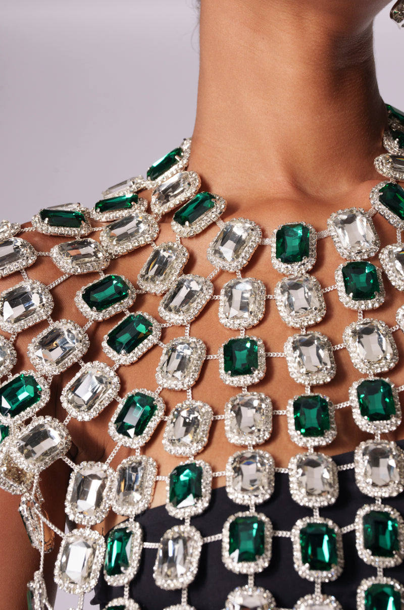 A close-up showcases an individual in a BEJEWELED EMERALD SLEEVELESS TOP, featuring interlinked large rectangular gems in green and clear, set against a black background. These sparkling metallic-framed gemstones form a luxurious and eye-catching pattern.