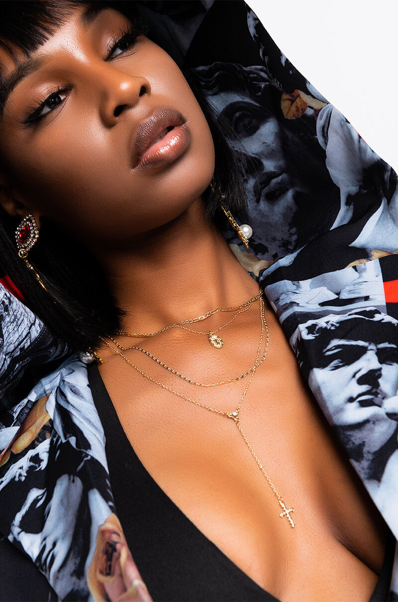 A woman with short black hair and glossy lips wears a black top and the BEST OF LUCK LONG LAYERED NECKLACE by Y&S Davidson Inc, which features a cross pendant. She also has on a patterned jacket adorned with artistic prints of faces and dramatic expressions. She looks away from the camera.