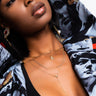 A woman with short black hair and glossy lips wears a black top and the BEST OF LUCK LONG LAYERED NECKLACE by Y&S Davidson Inc, which features a cross pendant. She also has on a patterned jacket adorned with artistic prints of faces and dramatic expressions. She looks away from the camera.