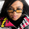 A person with long dark hair partially covering their face is wearing the BETTER LUCK YELLOW LENS SUNNIES and a colorful graphic leather jacket. The wind is blowing strands of hair across their face, and they have a neutral expression.