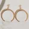 A pair of "BETTER THAN YOUR AVERAGE EARRING," gold crescent moon-shaped earrings adorned with small, sparkling crystals, are laid on a light-colored surface. Each earring features a small star at the top, also embellished with crystals, connecting to the crescent shapes.