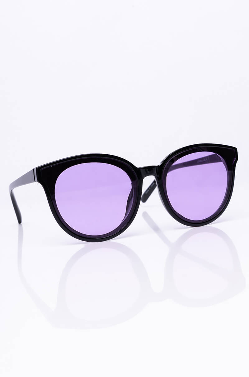 The BETTY ROUND SUNNIES, featuring black frames with round, purple-tinted lenses, are showcased against a white background. These sunglasses boast thick arms and a glossy finish, with a subtle reflection on the surface.