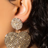 Close-up of an earlobe adorned with the BIG HEARTED RHINESTONE EARRING. This luxurious accessory features a large, sparkling round stud connected to a heart-shaped pendant. Both the stud and pendant are encrusted with numerous small, shiny crystals, creating an exquisitely glamorous look.