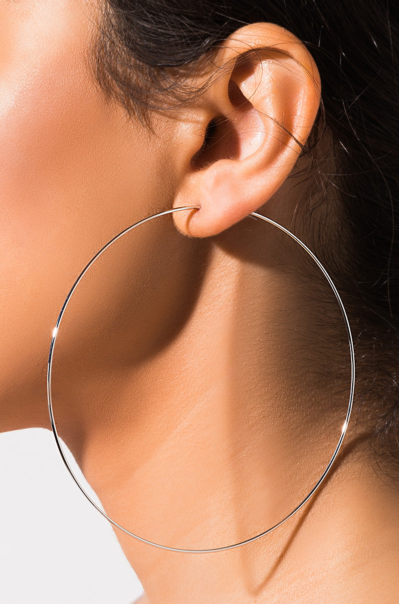 A close-up shot showcases a woman's ear adorned with the SEXY HOOP EARRING by MONTANA. The large, thin, gold hoop provides a striking contrast to her skin tone as it gracefully rests against her neck. The minimal background accentuates the intricate details of both the earring and her skin.