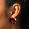 Close-up of a person’s ear adorned with the BIG RED EARRING, which features a round, deep red gemstone set in a gold-tone frame. The person has dark hair pulled back, allowing the earring to stand out prominently against their skin.