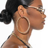 Profile view of a woman with smooth, pulled-back hair in a bun, wearing stylish glasses with a zigzag detail, BIGGER AND BETTER OVERSIZED HOOPS encrusted with stones, and a matching gold chain necklace. She is dressed in a black top.