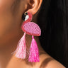 Close-up of a person wearing the BILLIE FLAMINGO EARRINGS, featuring two striking beaded oval shapes in pink and red, each adorned with pink and white tassels. The person has dark hair pulled back and is wearing a black strap.