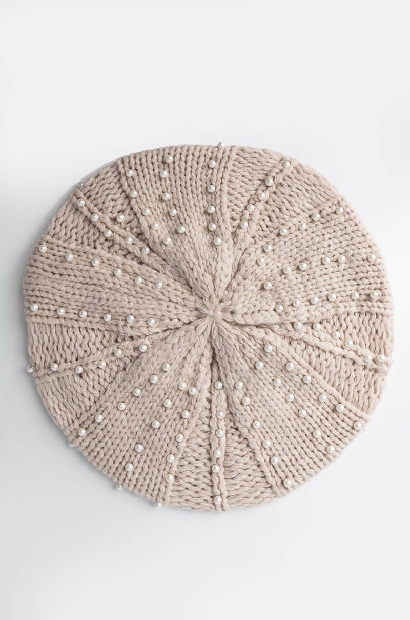 The BILLIE PEARL KNIT HAT, a beige knitted beret, is adorned with small white pearls evenly distributed across its surface in a radial pattern. Its textured yarn and delicate embellishments give it a stylish and elegant appearance against the plain white background.