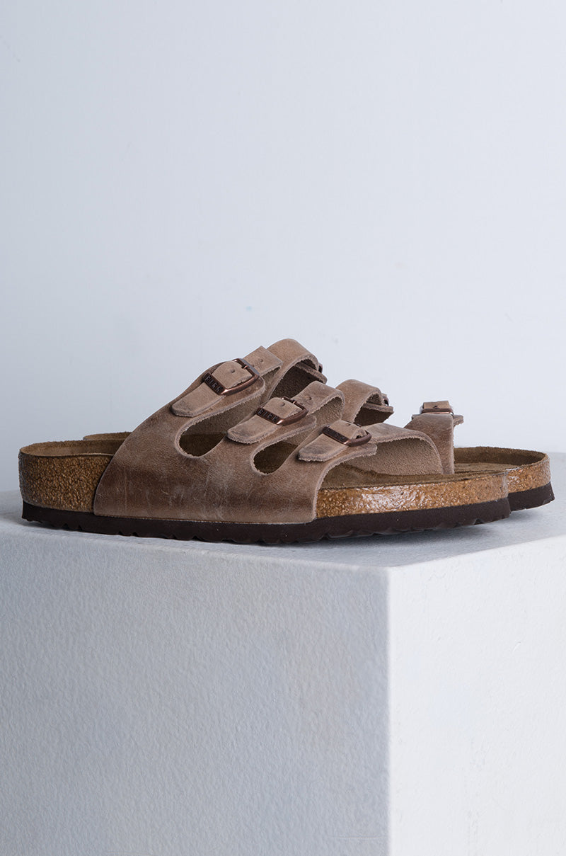 A pair of brown, suede SEXY CASUAL FLAT sandals featuring multiple adjustable straps, cork-latex footbeds, metal buckles, and dark soles. They are set on a white platform against a plain background.