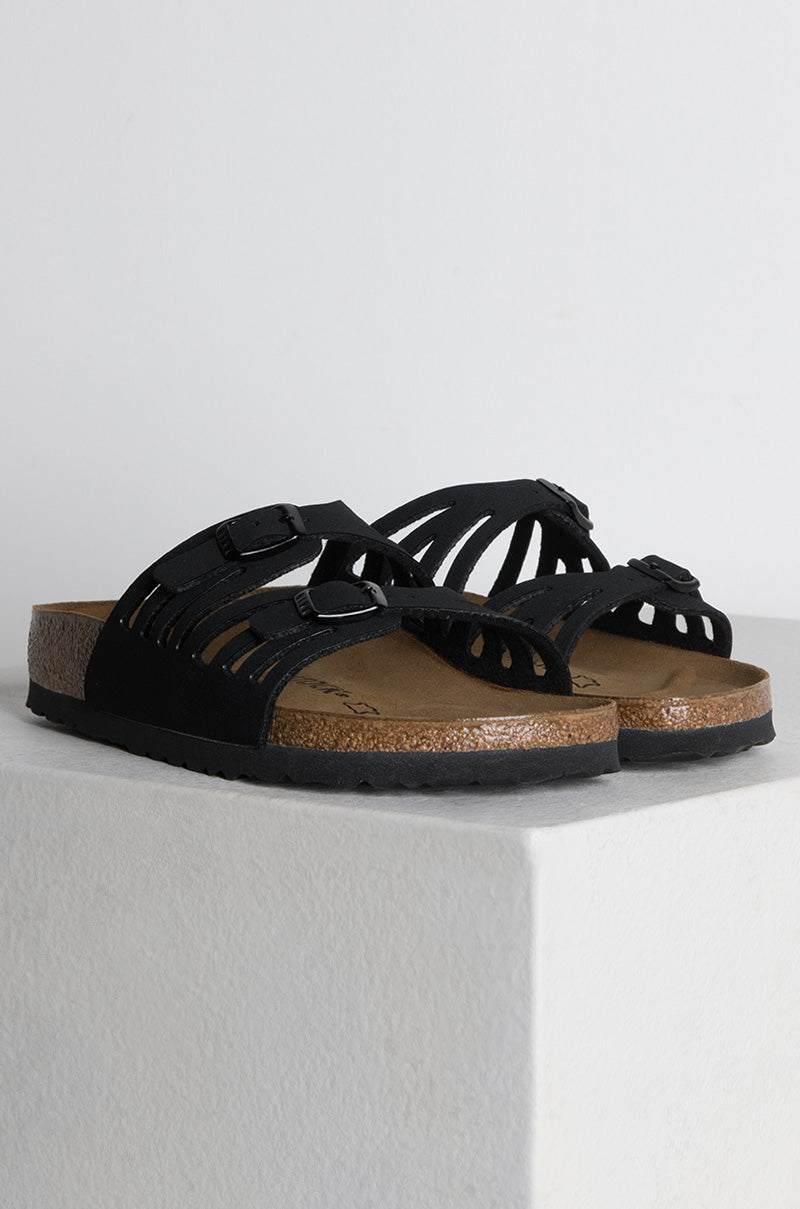 The SEXY CASUAL FLAT, a pair of black open-toe sandals with multiple slim straps, each featuring a small adjustable buckle, is displayed on a white surface. The sandals boast a cork footbed and a black rubber sole.