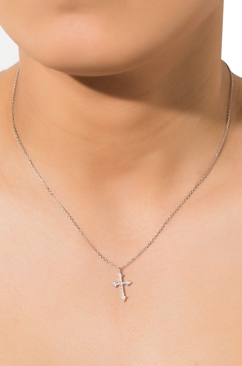 A close-up of a person wearing the BLESSINGS NECKLACE, a delicate silver chain featuring a sparkling cross-shaped pendant adorned with small, embedded gems that add a touch of shine. The background is plain white, and the person's face is partially visible.