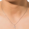 A close-up of a person wearing the BLESSINGS NECKLACE, a delicate silver chain featuring a sparkling cross-shaped pendant adorned with small, embedded gems that add a touch of shine. The background is plain white, and the person's face is partially visible.