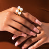 Close-up of two hands with long, manicured nails painted in a pale pink polish. One hand holds the other gently, showcasing the BLOOM PEARL COCKTAIL RING adorned with three large pearls. The person wears a brown fabric that contrasts beautifully with the hands and ring.