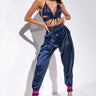 A woman poses confidently wearing the stylish BODY PARTY JOGGER in a midnight blue satin ensemble. Her outfit features a strappy crop top paired with high-waisted, loose-fitting joggers. She accessorizes with a necklace and bright pink high-heeled sandals against a minimal white background.