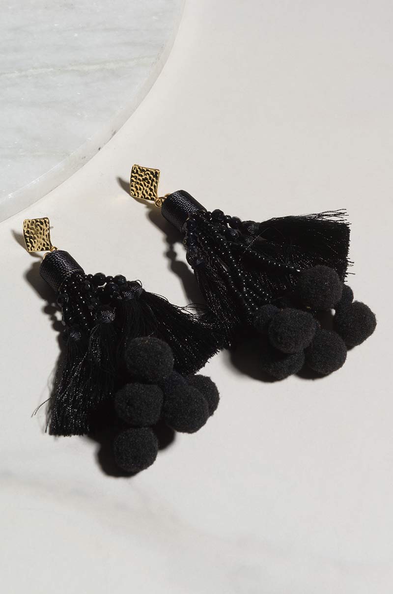 A pair of SEXY DANGLE EARRING, featuring black cords hanging in a tassel style with small black pom-poms clustered at the ends and beads at the top, all connected to gold stud bases. They are displayed on a white marble and light grey surface.
