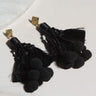 A pair of SEXY DANGLE EARRING, featuring black cords hanging in a tassel style with small black pom-poms clustered at the ends and beads at the top, all connected to gold stud bases. They are displayed on a white marble and light grey surface.