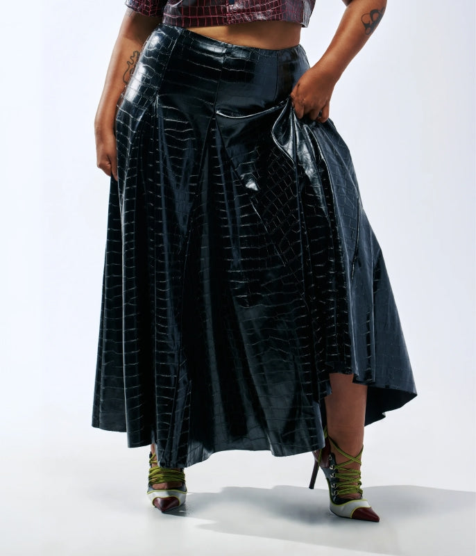 A model wearing a high-waisted, black faux-croc-textured midi skirt with a draped side detail, paired with green lace-up heels, shot against a white studio background.