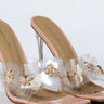 Introducing the BUTTERFLY SECRET CLEAR MULE: a pair of elegant high-heeled shoes with clear straps featuring delicate floral decorations adorned with small gold and rhinestone embellishments. The heels are transparent, and the insoles have a neutral beige color.