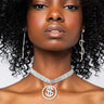 A woman with curly black hair wears the CA CHING! DOLLAR SIGN CHOKER and sparkling, dangling earrings. She has a calm expression and is posed against a plain white background.