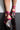 The image features a close-up of someone wearing the MAMA CAN I GET AN AMEN CHUNKY STRAPPY SANDAL, which has pink strappy high heels with clear, wedge-shaped heels. The individual stands on a glossy black surface against a plain white background, showcasing the intricate straps that wrap around their ankles and feet.