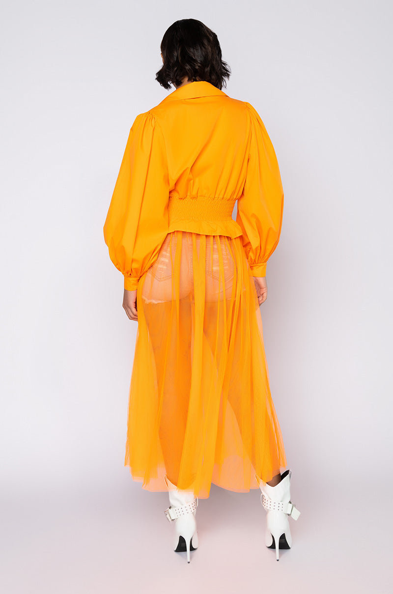 A model stands against a plain background, wearing the CAN'T FOOL ME MAXI DRESS WITH TULLE in vibrant orange, featuring long sleeves and button-up details. The dress includes a matching sheer, floor-length skirt. They sport white high-heeled shoes adorned with bow details on the ankle straps. The model is facing away from the camera.