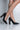 A close-up of a pair of legs wearing the "CANT HAVE JUST ONE CHUNKY HEEL POINTED TOE PUMPS," featuring black high-heeled shoes with glittering details on the block heels, standing against a plain, light-colored background. The image highlights the stylish design of this chic footwear.