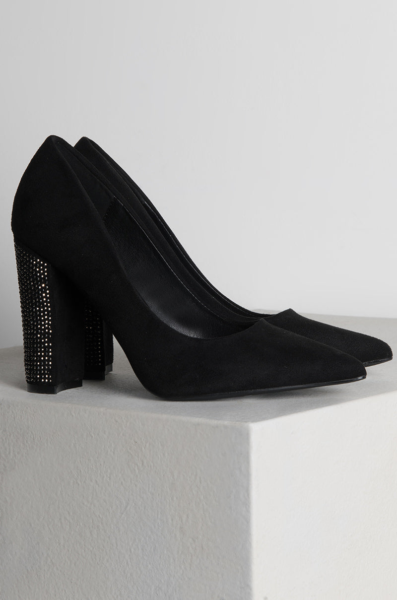 The CANT HAVE JUST ONE CHUNKY HEEL POINTED TOE PUMPS are elegantly displayed on a white platform. These black high-heeled shoes feature sparkling rhinestones on the heels, adding a touch of glamour to their sleek, matte finish. The pointed toes and sturdy block heels enhance their chic design.