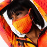 A person with short black hair is wearing a vibrant orange and red textured CAN'T STOP WON'T STOP MASK outfit that has a zip-up front. The individual has their arms raised, touching their head, revealing hoop earrings visible through their bob haircut.