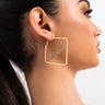 Close-up of a person with dark hair pulled back, showcasing the CAPRICORN PAVE HOOP earrings. The large square hoops are gold with a script design in the center. The individual has small piercings on the ear and is looking to the side against a neutral background.