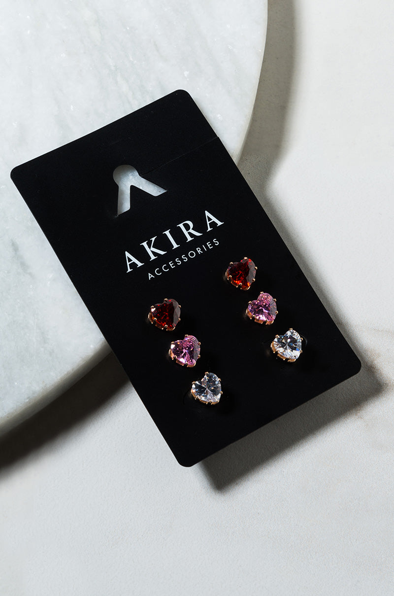 A set of CARRIE EARRING SET stud earrings on a black display card, featuring three pairs: one with red gemstones, one with pink gemstones, and one with clear gemstones, all in a shiny, reflective finish. The card is placed on a light-colored, textured surface.