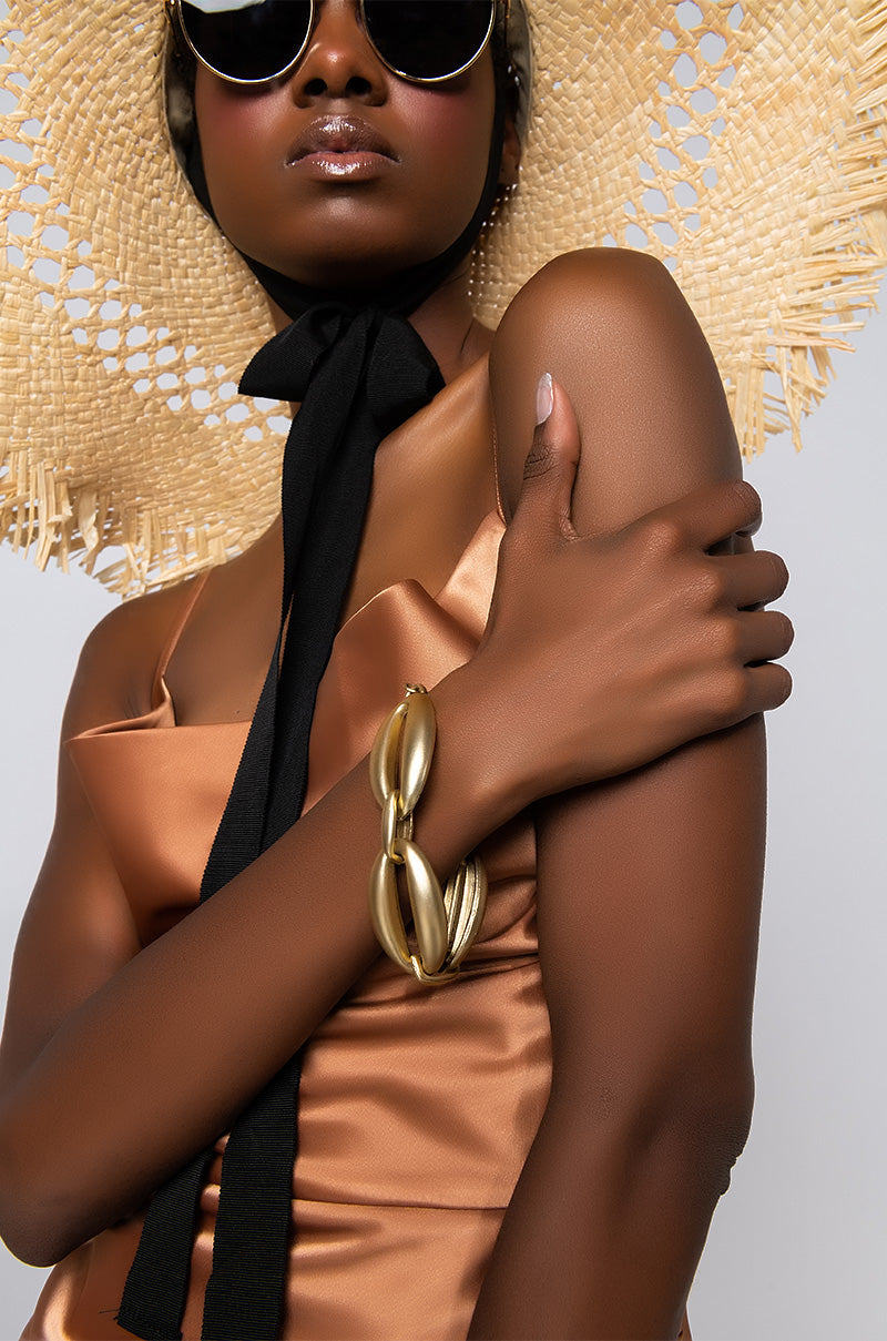 A woman wearing a large straw hat with a black ribbon, dark sunglasses, and a sleeveless bronze satin dress stands with her arms crossed, showcasing the BIG LING BRACELET on her wrist. The plain background accentuates her fashion accessories.