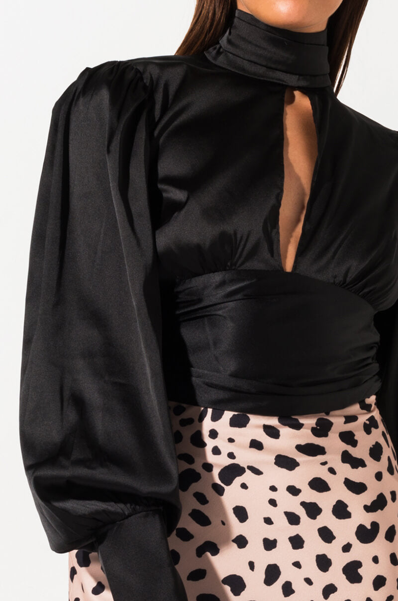 A woman wearing the CHECKMATE LONG SLEEVE BLOUSE, a black satin top with a high neck and puffy long sleeves, featuring a keyhole cutout at the chest. She is also wearing a light-colored skirt with black polka dots.