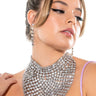 A woman with tied-back hair and winged eyeliner looks downwards, elegantly adorned in the CHEST ARMOR NECKLACE SET featuring a glamorous, large, intricate diamond necklace and matching long earrings. She is dressed in a light purple, sleeveless top or dress against a plain white background.