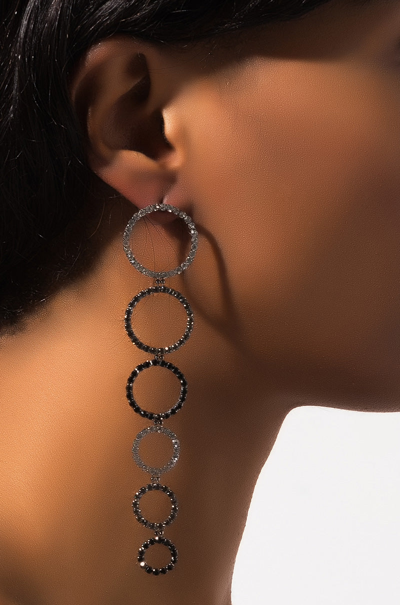 A close-up view of a person wearing the EARRING featuring five interconnected circles. These circles, which increase in size from bottom to top, are adorned with small, sparkling stones. The background is blurred to keep the focus on the EARRING and the person's neck and ear.