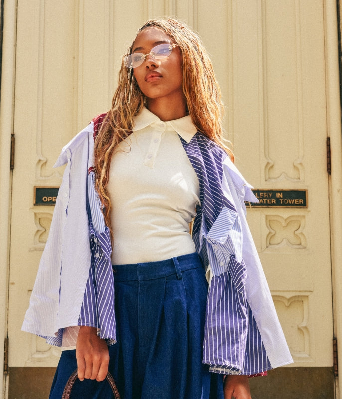A woman wearing a white polo shirt, a white shirt with purple and blue stripes draped over her shoulders, and a dark blue skirt, standing in front of a cream-colored building door.