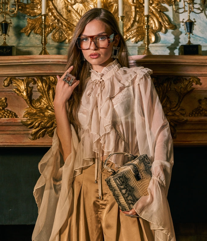 A woman models a sheer champagne beige ruffled blouse with a high neck and bow tie detail, paired with a tan skirt. She is holding a textured black and beige handbag and wearing distinctive pink-tinted square eyeglasses, posed in front of an ornate gold fireplace.