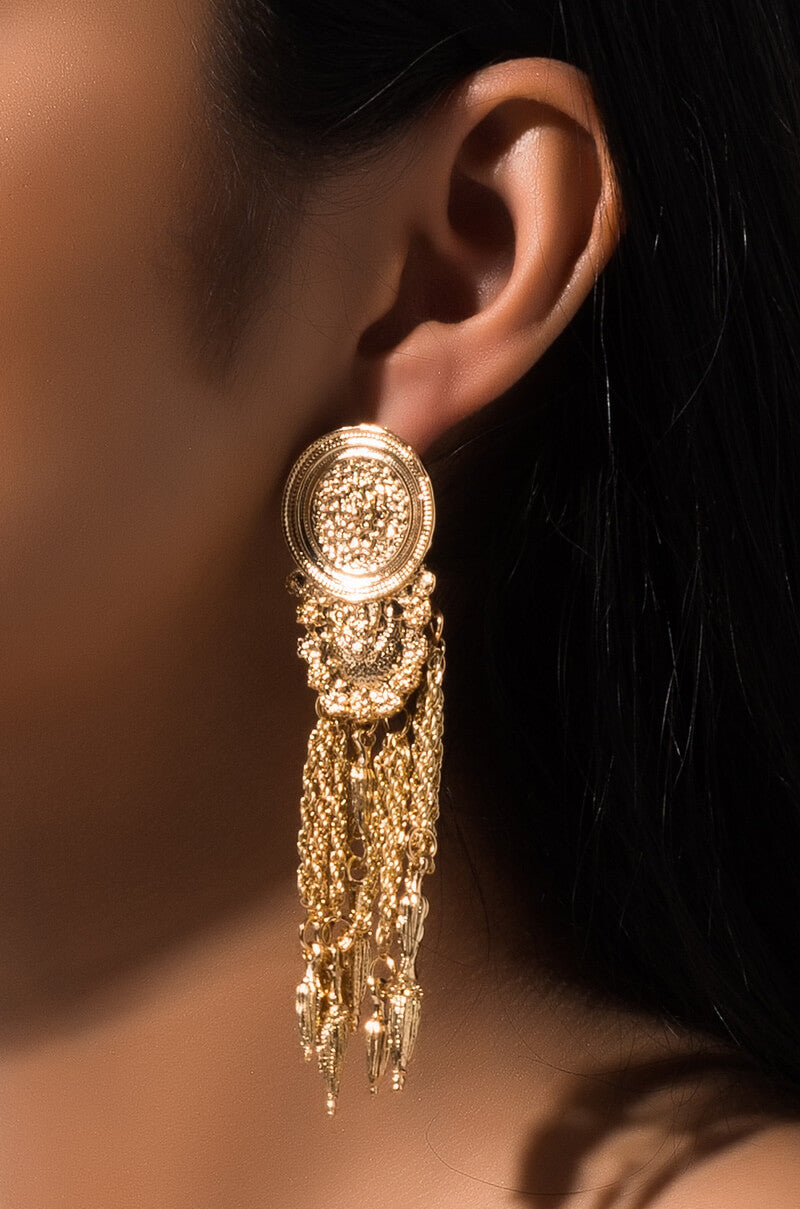 Close-up of a person's ear adorned with the stylish CLUB ROYALE DANGLE EARRING. This exquisite piece features a circular stud with intricate, elaborate designs and multiple dangling gold chains, adding an elegant and luxurious touch to the overall aesthetic.