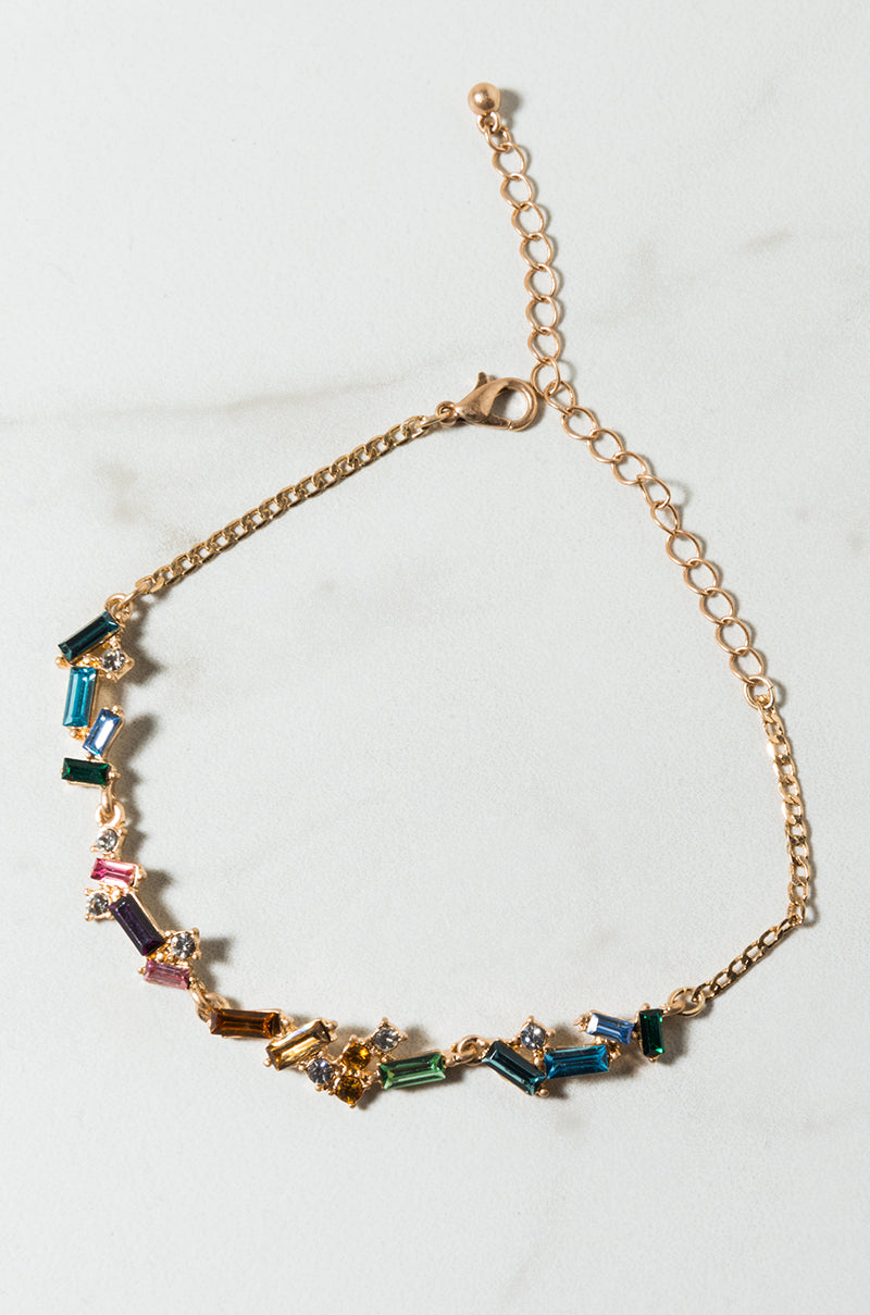 The COLOR RUSH LINK BRACELET features a golden chain adorned with multicolored, rectangular gemstones arranged in a captivating pattern. The adjustable chain includes links and a small clasp for secure fastening. It is elegantly displayed on a light, textured surface.