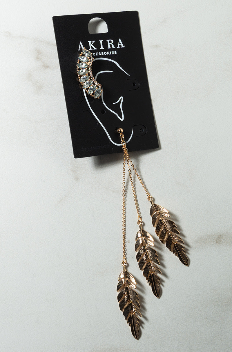 The DROP IT NOW FEATHER EAR SET from AKIRA Accessories is showcased against a light marble background. The ear cuff features rhinestones and is connected to three dangling gold feather charms by intricate gold chains. The sophisticated piece of jewelry is presented on its sleek black packaging card.
