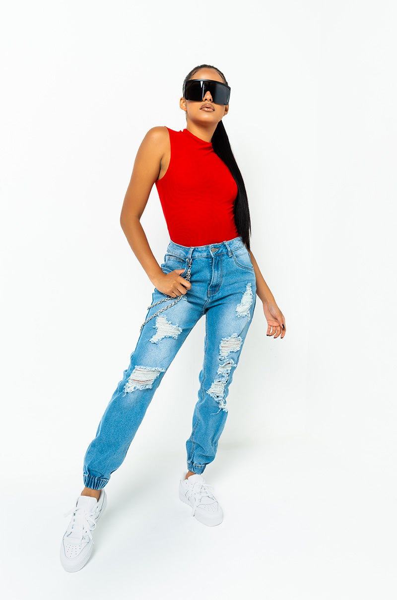 A person with long black hair stands confidently wearing a red sleeveless top, COMFY AND CUTE BOYFRIEND JEANS in distressed blue, and white sneakers. They have oversized black sunglasses and one hand in their pocket, striking a pose against a plain white background.