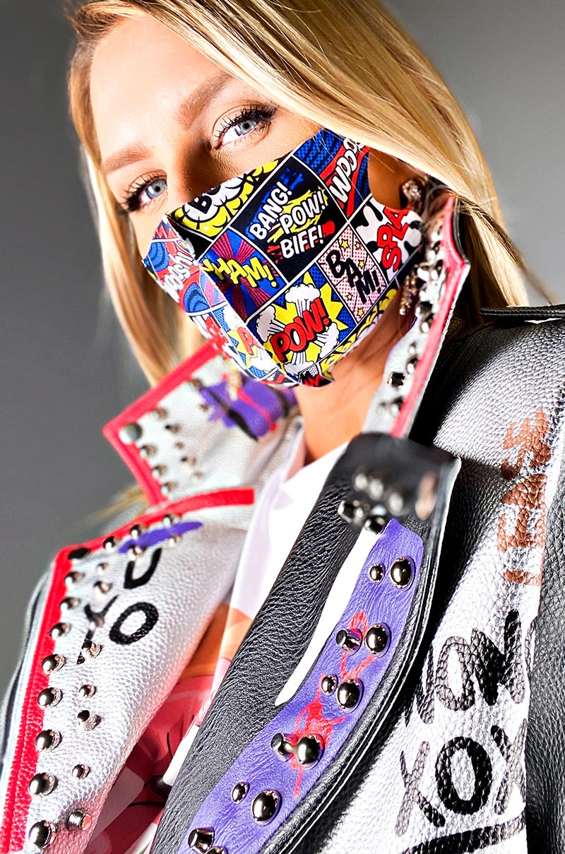 A person with long blonde hair wears a colorful COMIC RELIEF FASHION MASK COVER and a studded leather jacket adorned with graffiti-style writings and patches. The jacket features a mix of pop-art elements, including bold text and vibrant patterns, all set against a neutral background.