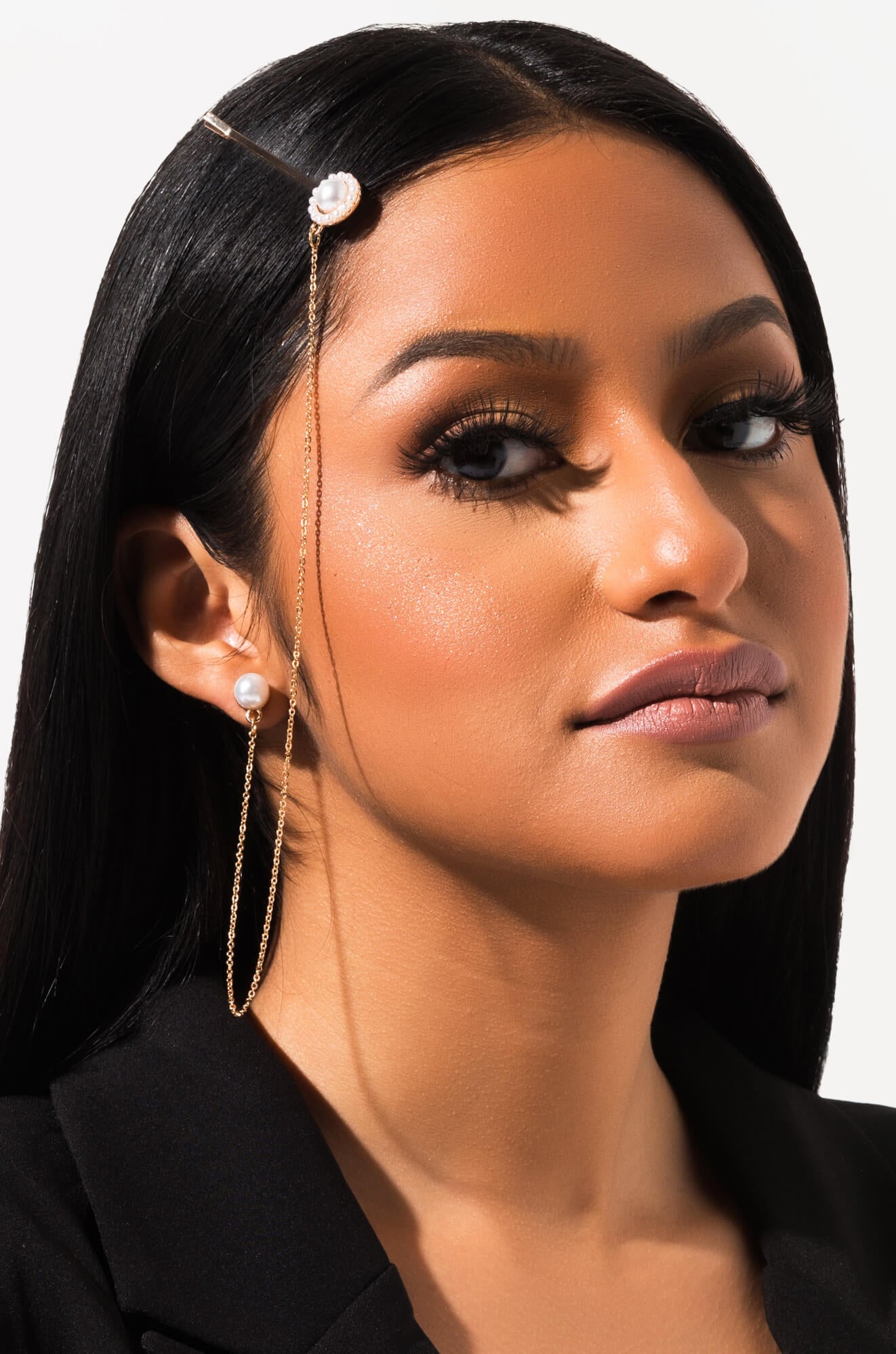 A close-up of a woman with long, straight black hair. She is wearing a thin gold head chain with a pearl accent and matching HAIR PIN EARRINGS, which are long gold earrings adorned with pearls. Her makeup is glamorous with long lashes and nude lipstick. She is dressed in a black top or jacket.