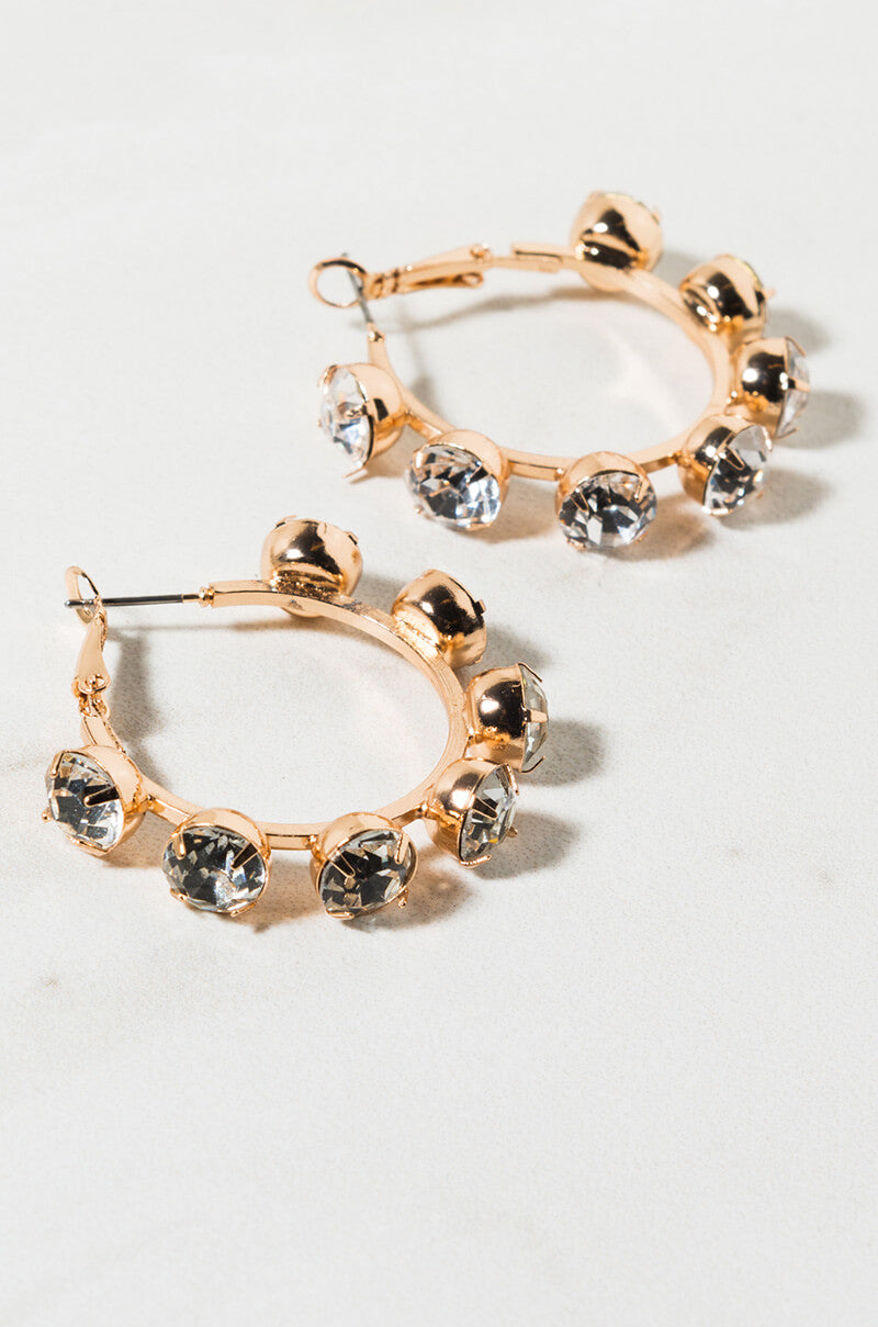 The TRUBULA RHINESTONE HOOP EARRING features a pair of gold hoops accented with large, evenly spaced clear rhinestones. The gemstones are meticulously distributed around the hoops, creating a bold and elegant look. These earrings are showcased on a light-colored surface.