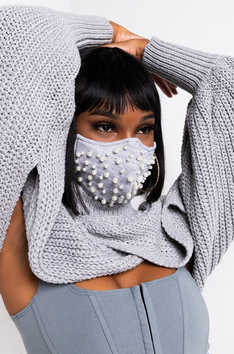 A person with straight black hair is wearing a light gray sweater and a matching COZY AND CLASSY PEARL DETAIL FASHION FACE COVER. The person's right arm is raised, holding the back of their head. The background is plain white.